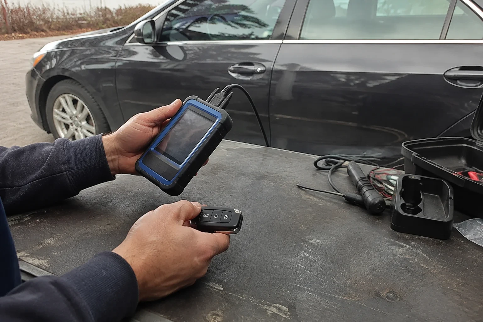 Hands holding a blue-edged electronic device and car-key fob at a worktable beside a parked car.